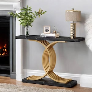 Puede incluir: Una mesa consola de mármol negro con una base de metal dorado. La base presenta un diseño curvo en forma de X. La mesa está decorada con un pequeño jarrón de vegetación, un libro y una foto enmarcada.