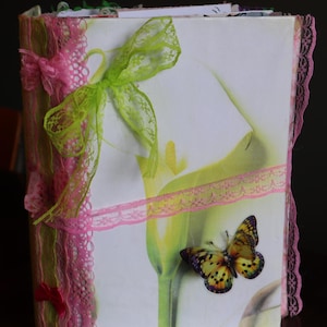 May include: A rectangular journal featuring a white cover with a large green and white calla lily illustration and a yellow and black butterfly. The spine and edges are decorated with pink lace trim, a bright green ribbon bow, and small pink bows.