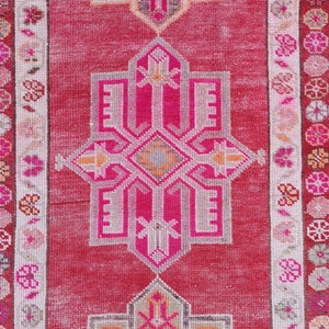 May include: A vintage Turkish runner rug with a pink and white geometric pattern. The rug has a faded, distressed look and features a central medallion design.