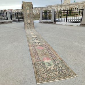 May include: A long, rectangular rug with a beige background and intricate patterns in shades of brown, green, pink, and white. The rug is laid out on a concrete surface.