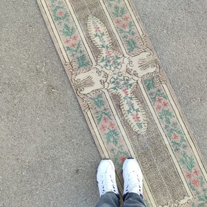 May include: A rectangular, vintage-style rug with a floral pattern in shades of pink, green, and beige. The rug has a central design with a border featuring stripes and floral motifs. The rug is placed on a concrete surface.