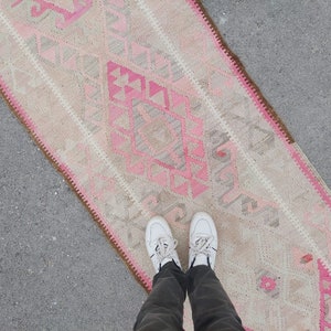 May include: A pink and white patterned rug with a brown fringe. The rug is laid out on a gray concrete surface.