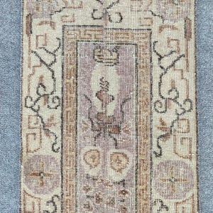 May include: A vintage runner rug with a light beige background and a geometric pattern in shades of brown, black, and pink. The rug has a fringed edge.