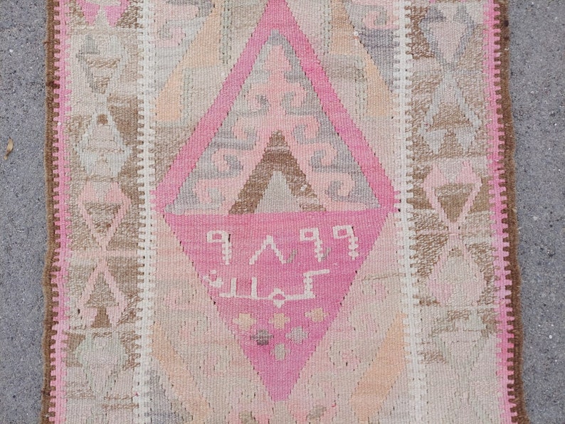 May include: A pink and brown patterned rug with a diamond-shaped design in the center. The rug has a fringe border and features a geometric pattern with Arabic script.