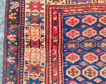Op de afbeelding: Close-up van een gedeelte van een vintage tapijt met een gedetailleerd geometrisch patroon. Het tapijt heeft een diepblauw veld met rode en bruine accenten, omlijst door randen in rood, beige en teal. Het ontwerp bevat herhalende vormen en bloemmotieven.