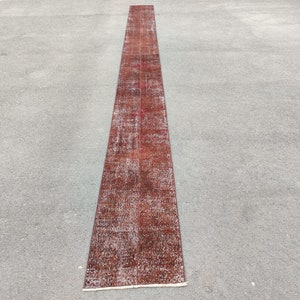 Peut inclure: Un tapis vintage long et étroit, rouge et brun, à l'aspect vieilli. Le tapis est posé à plat sur une surface en béton gris.