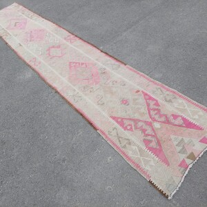 May include: A long, narrow rug with a pink and white geometric pattern. The rug has a fringe on both ends.