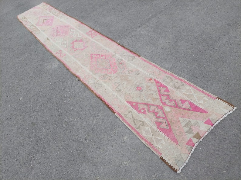 May include: A long, narrow rug with a pink and white geometric pattern. The rug has a fringe on both ends.