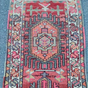 May include: A rectangular woven rug featuring a central diamond medallion in shades of coral, pink, and charcoal gray. The border displays a repeating geometric pattern in cream, pink, and gray, with a textured pile and fringed edges.
