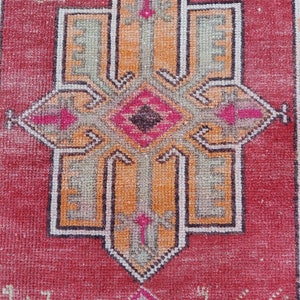 May include: A vintage Turkish rug with a red background and a central design featuring a cross with intricate details in shades of orange, green, and black.