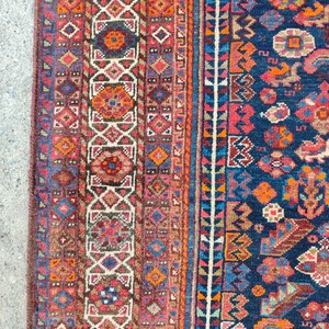 May include: A vintage rug with a colorful geometric pattern. The rug has a dark blue background with red, orange, and white accents. The rug is woven with intricate details and has a worn, antique look.