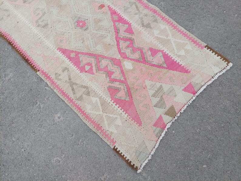 May include: A pink and beige patterned runner rug with fringe on the edges. The rug has a geometric design with a diamond pattern.