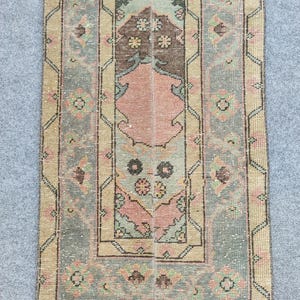 Pode incluir: Um tapete retangular vintage com um design central em tons de rosa, castanho e verde. O tapete apresenta um padrão floral detalhado com uma borda bege e uma aparência desbotada e desgastada, sugerindo idade e uso. As dimensões do tapete não são visíveis.