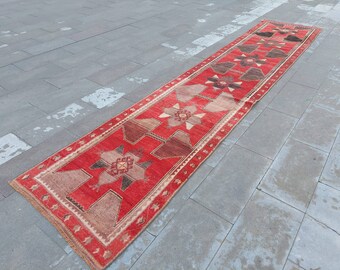 Puede incluir: Una alfombra de pasillo roja, marrón y beige con un patrón geométrico. La alfombra tiene un borde con flecos y está colocada sobre un suelo de baldosas grises.
