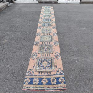 May include: A long, narrow rug with a pink background and a blue and cream geometric pattern. The rug has a fringe on both ends.