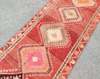 May include: A red, pink, and beige runner rug with a geometric pattern. The rug has a fringe border and is made of wool.