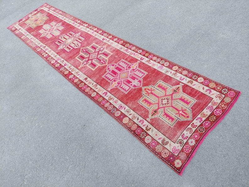May include: A long, narrow red runner rug with a white border and intricate pink and beige geometric patterns.