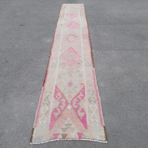 May include: A long, narrow rug with a pink and white geometric pattern. The rug has a fringe on both ends.