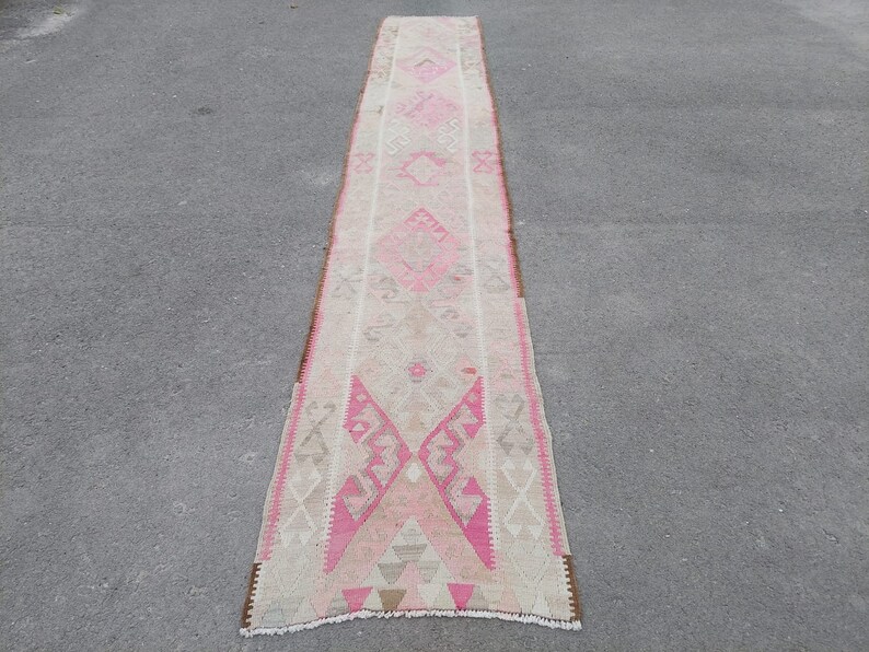May include: A long, narrow rug with a pink and white geometric pattern. The rug has a fringe on both ends.