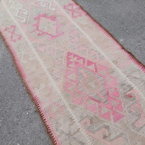May include: A long, narrow rug with a pink and beige geometric pattern. The rug has a fringe border.