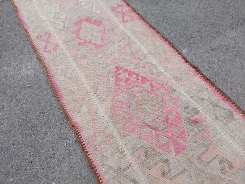 May include: A long, narrow rug with a pink and beige geometric pattern. The rug has a fringe border.