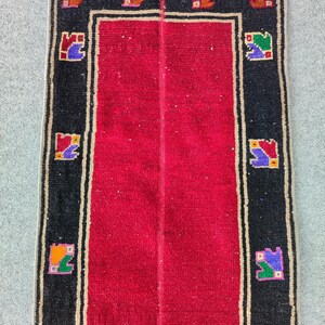 May include: A rectangular rug with a red centre and a black border. The border features a repeating pattern of colourful geometric shapes. The rug has a beige fringe on one end.