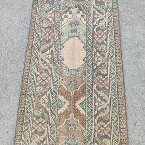 May include: Rectangular vintage rug with a central design in beige, brown, and teal. The rug features intricate floral and geometric patterns, with a border in shades of gray and beige. The rug is likely made of woven material.