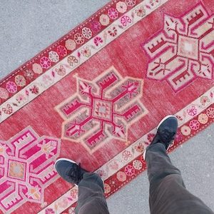 May include: A red and pink runner rug with a geometric pattern. The rug has a fringed border and is made of a soft, woven material.