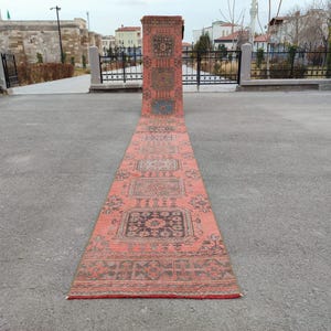 Puede incluir: Una alfombra vintage larga y rectangular con una combinación de colores rojo y marrón descoloridos. La alfombra presenta intrincados patrones geométricos y motivos florales. La alfombra se exhibe al aire libre sobre una superficie gris.