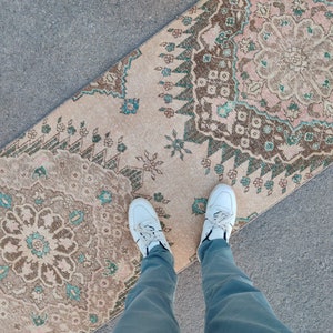 May include: A beige and brown patterned rug with green accents. The rug is laid out on a gray surface. A person is standing on the rug wearing white sneakers and blue jeans.