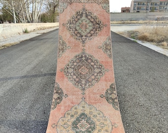 May include: A rectangular, vintage-style rug with a faded, distressed appearance. The rug features a salmon-coloured background with intricate, repeating patterns in shades of brown, green, and cream. The rug is displayed outdoors on an asphalt surface.