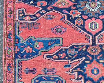 May include: Close-up of a section of an antique rug with a detailed, intricate pattern. The rug features a central field of coral-red, framed by a border of dark blue and accents of pink, green, and cream. The design includes floral and geometric motifs.