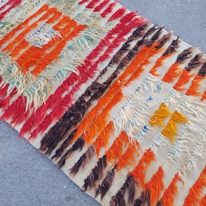 May include: A white, handwoven rug with a geometric pattern in shades of orange, red, brown, and blue. The rug has a shaggy texture and is made with long, loose fibers.