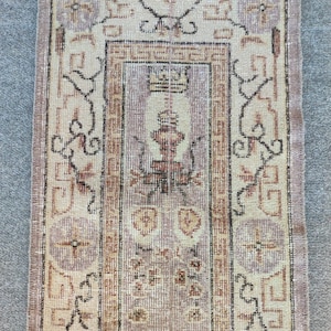 May include: A vintage rug with a light brown background and a central design featuring a stylized floral motif. The rug has a border with a repeating geometric pattern in shades of brown and black.