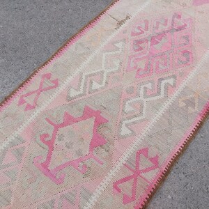 May include: A pink and beige runner rug with a geometric pattern. The rug has a brown fringe border.