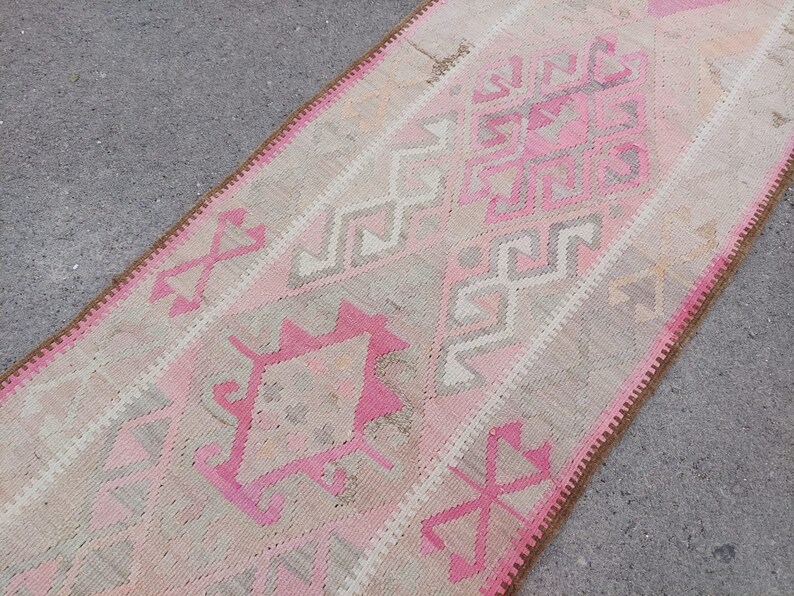 May include: A pink and beige runner rug with a geometric pattern. The rug has a brown fringe border.
