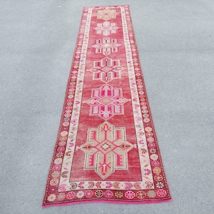 May include: A long, narrow red runner rug with a pink and white geometric pattern. The rug has a fringe on both ends.