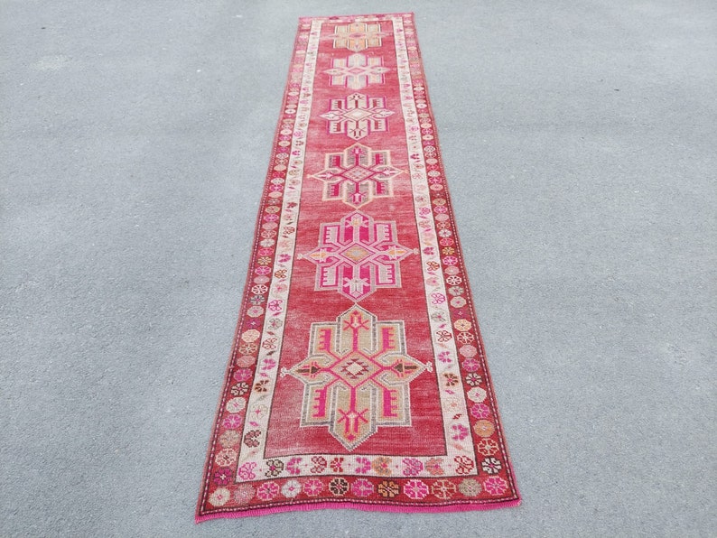 May include: A long, narrow red runner rug with a pink and white geometric pattern. The rug has a fringe on both ends.
