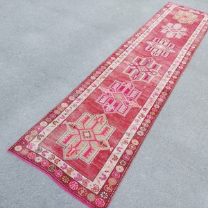 May include: A long, red runner rug with a traditional Turkish design. The rug features intricate geometric patterns in shades of pink, beige, and white.