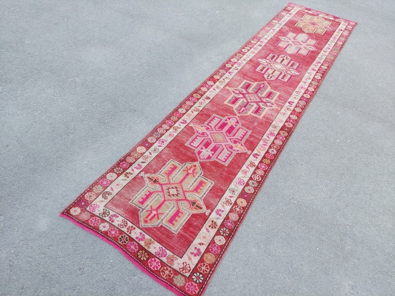 May include: A long, red runner rug with a traditional Turkish design. The rug features intricate geometric patterns in shades of pink, beige, and white.
