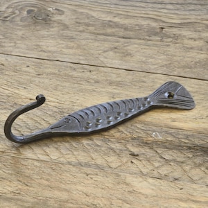 May include: A handcrafted, dark metal fish hook. The hook is shaped like a fish, with a curved hook at one end and a tail fin at the other. The fish hook is approximately 8 inches long and is designed to be mounted on a wall.