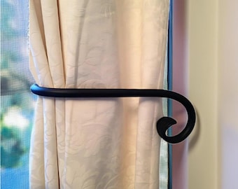 May include: Black metal curtain tieback with a decorative scroll design. The tieback is holding back a white curtain.