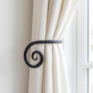 May include: A black metal curtain holdback featuring a hammered, textured finish and a prominent spiral design. The holdback is mounted to secure a long, off-white fabric curtain panel, keeping the fabric gathered neatly to one side of the window.