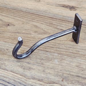 Puede incluir: Un gancho de pared de metal gris oscuro con un diseño curvo y una placa de montaje rectangular con dos orificios pretaladrados. El gancho tiene un acabado metálico liso y un extremo ligeramente cónico para colgar objetos.