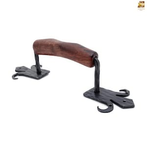 Op de afbeelding: Handgesmede ijzeren deurgreep met een donkerhouten handvat. De zwarte ijzeren basis heeft een getextureerde afwerking en decoratieve rondingen. Een rustiek, handgemaakt stuk voor deuren, kasten of laden.