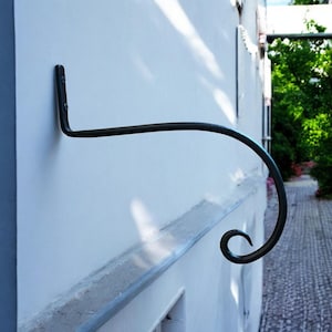 Könnte beinhalten: Schwarzer Wandhaken aus Metall mit dekorativem Schnörkeldesign. Der Haken ist an einer weißen Wand montiert.