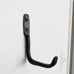 May include: A matte black wall hook with a textured, hand-forged appearance. The hook features a vertical mounting plate secured to the wall with two black Phillips head screws, extending outward into a curved J-shaped arm for hanging items.