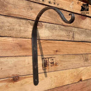May include: A black metal wall-mounted hook with a curved design. The hook is attached to a wooden wall.