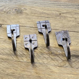 Puede incluir: Cuatro ganchos de pared artesanales de metal gris oscuro con varios diseños decorativos en la parte superior. Cada gancho tiene una sección inferior curvada para colgar objetos. Los ganchos están dispuestos sobre una superficie de madera rústica.