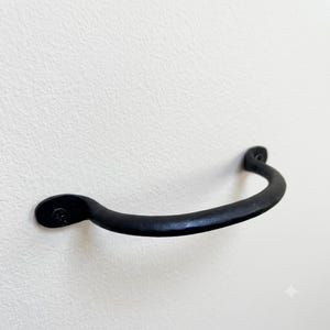 May include: A matte black metal drawer pull with a curved, arched design. The handle features a textured, hand-forged appearance with two circular mounting points at each end, each secured by a single visible screw head on the flat base.